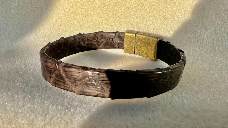 Pu&ograve; includere: Un braccialetto in pelle marrone con un motivo strutturato a forma di serpente. Il braccialetto presenta una chiusura magnetica rettangolare color oro. Il braccialetto &egrave; largo circa 2 cm.