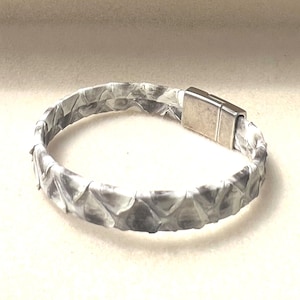 Peut inclure: Un bracelet à motifs gris et blancs avec un fermoir magnétique argenté. Le bracelet est fait d'une matière texturée, peut-être du cuir ou un tissu similaire. Le fermoir est rectangulaire et semble être en métal.
