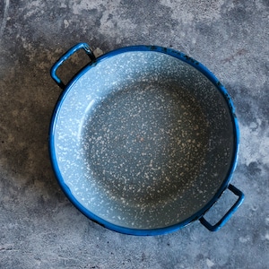 Op de afbeelding: Een blauwe emaille pan met een gespikkelde witte binnenkant en twee handvatten. De pan staat op een grijs betonnen oppervlak.