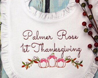 Könnte beinhalten: Weißes Baby-Lätzchen mit Rüschenkante, bestickt mit "Palmer Rose 1st Thanksgiving" in bordeauxfarbenem Faden. Das Design umfasst drei Kürbisse mit rosa Schleifen und Herbstblättern. Rechts ist ein dekorativer Stiel mit roten und grünen Beeren.