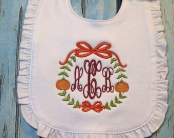 Könnte beinhalten: Weißer Baby-Lätzchen mit einem Rüschenrand und einem gestickten Design mit einem Monogramm, das von Kürbissen und Blättern umgeben ist. Das Monogramm ist in Burgunderrot und die Kürbisse und Blätter sind in Orange- und Grüntönen.