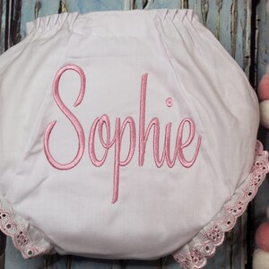 May include: White baby bloomers with a pink embroidered name "Sophie" and lace trim.