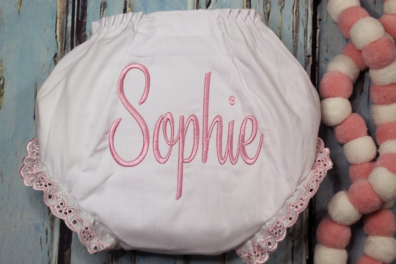 May include: White baby bloomers with a pink embroidered name "Sophie" and lace trim.