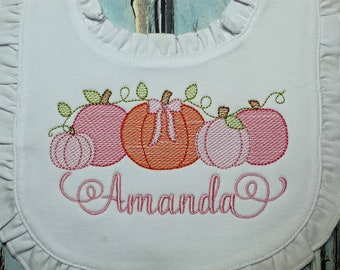 Könnte beinhalten: Eine weiße Lätzchen mit einem gestickten Kürbisdesign in Rosa und Orange und dem Namen "Amanda" in rosa Kursivschrift.