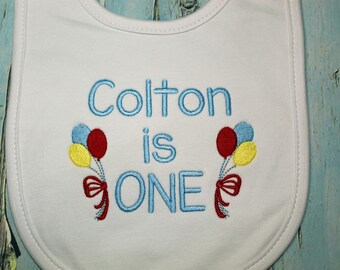 May include: White baby bib embroidered with the words "Colton is ONE" in blue. The bib features two sets of balloons in red, yellow, and blue, tied with red bows. A celebratory design for a first birthday.