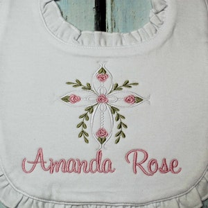 May include: White baby bib with a pink embroidered cross with floral accents and the name "Amanda Rose" embroidered below.