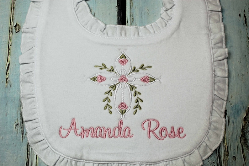 May include: White baby bib with a pink embroidered cross with floral accents and the name "Amanda Rose" embroidered below.