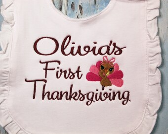 Könnte beinhalten: Weißer Lätzchen mit brauner Stickerei, auf dem "Olivia's First Thanksgiving" steht, mit einer Illustration eines rosa und braunen Truthahns.