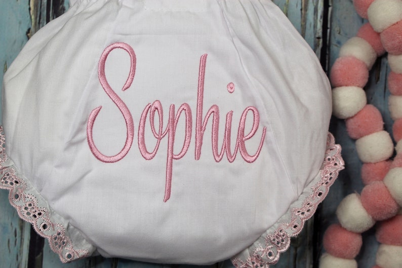 May include: White baby bloomers with pink lace trim and the name "Sophie" embroidered in pink.