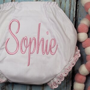 May include: White baby bloomers with pink lace trim and the name "Sophie" embroidered in pink.