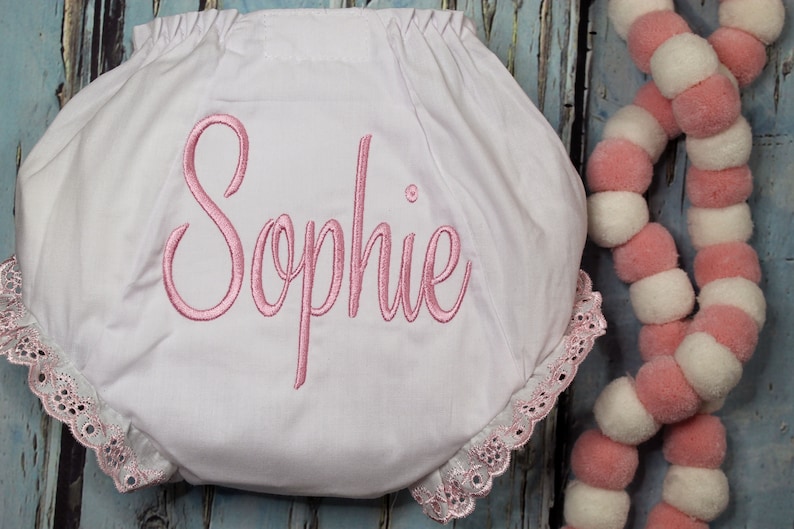 May include: White baby bloomers with pink lace trim and the name "Sophie" embroidered in pink.