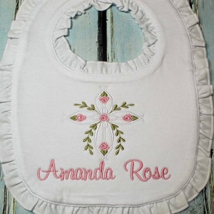 May include: White baby bib with a pink embroidered cross with pink roses and green leaves. The bib has a ruffled edge and the name "Amanda Rose" is embroidered below the cross.