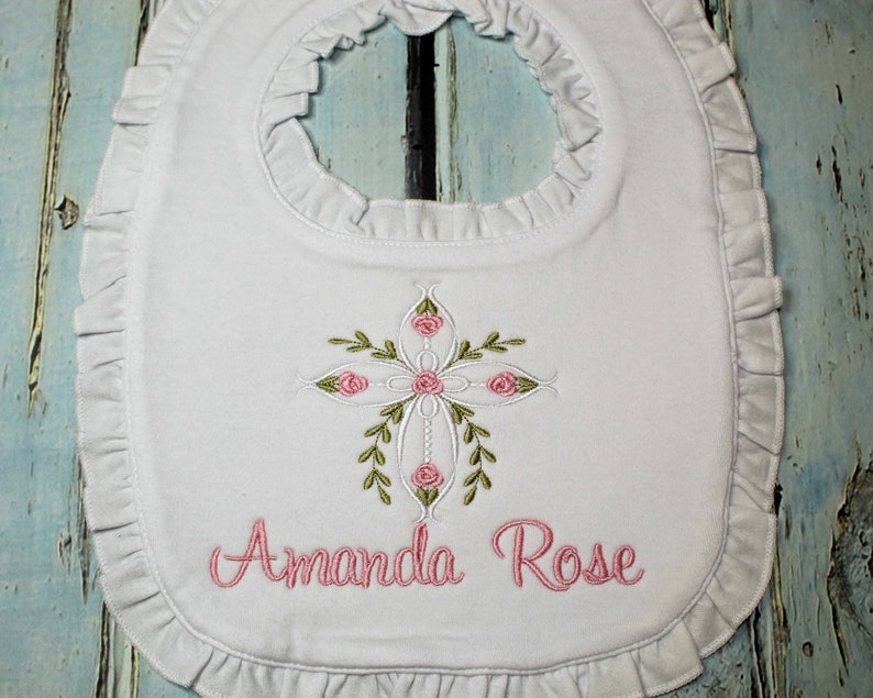 May include: White baby bib with a pink embroidered cross with pink roses and green leaves. The bib has a ruffled edge and the name "Amanda Rose" is embroidered below the cross.