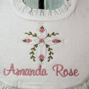 May include: White bib with a ruffled edge, featuring an embroidered cross with pink roses and green leaves. The name "Amanda Rose" is embroidered below the cross.