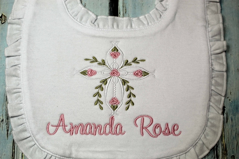 May include: White bib with a ruffled edge, featuring an embroidered cross with pink roses and green leaves. The name "Amanda Rose" is embroidered below the cross.