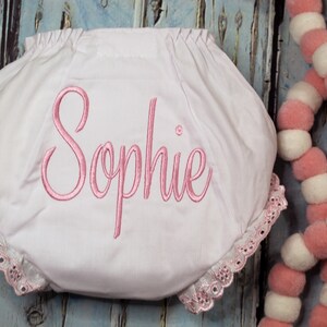 May include: White baby bloomers with pink lace trim and the name "Sophie" embroidered in pink.
