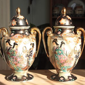 May include: Two ornate porcelain vases with gold accents and intricate floral and figure designs. The vases have a black and gold base and are decorated with a white background and colourful floral and figure designs.
