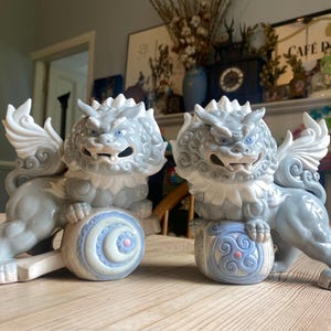 May include: A pair of ceramic guardian lion statues in light grey with white wings and collars. Each lion rests on a decorative base with blue and pink details. The statues have a fierce expression. The statues are decorative ornaments.