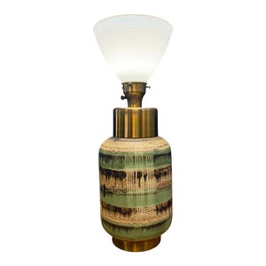 May include: A table lamp featuring a cylindrical ceramic base with horizontal bands of green, brown, and cream glaze. The lamp has a metallic gold-toned neck and a wide, flared, white glass shade with a subtle grid pattern on the exterior surface.