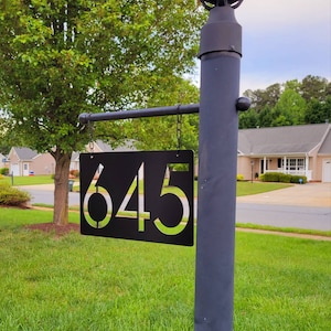 May include: Black metal address sign with the number 645 in a modern font. The sign hangs from a black metal pole with a decorative light fixture at the top. The sign is hanging in a grassy yard.