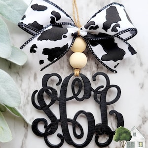 May include: Black wooden monogram with a cow print ribbon bow and wooden beads. The monogram is the letters "SGB".