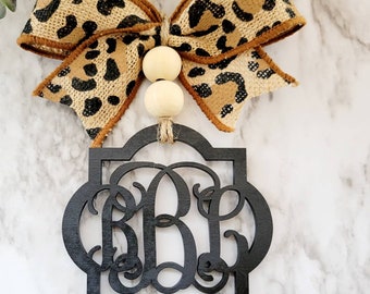 May include: Black wooden monogram with a leopard print bow and wooden beads. The monogram is in a square frame with a rounded top and bottom. The bow is tied with a brown ribbon.