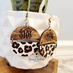 May include: A pair of wooden earrings with a leopard print design. The top half of each earring is a round piece of wood with a monogram engraved on it. The bottom half is a half-circle of leopard print fabric.