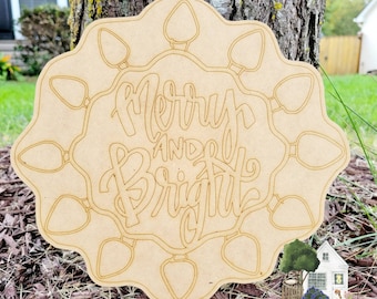 Puede incluir: Un recorte de madera de una corona de Navidad con el texto "Merry and Bright" en una fuente decorativa. La corona está decorada con una cadena de luces.
