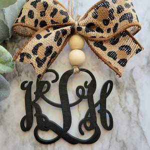 May include: A black wooden monogram ornament with the letters "K L H" in a cursive script. The ornament is decorated with a brown and black leopard print burlap bow and two wooden beads.