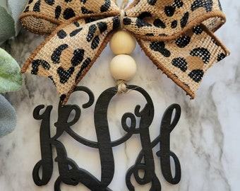 May include: A black wooden monogram ornament with the letters "K L H" in a cursive script. The ornament is decorated with a brown and black leopard print burlap bow and two wooden beads.