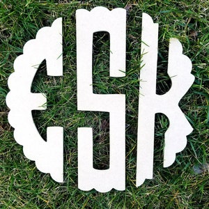 May include: A cream-colored wooden monogram with the letters CSK, set against a backdrop of green grass. The monogram is in a circular shape with a scalloped edge, creating a decorative and personalized design.