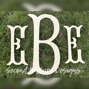 May include: Three wooden letters, E, B, and E, in a decorative font, are arranged in a row on a green grassy surface. The letters are unfinished and have a natural wood color.