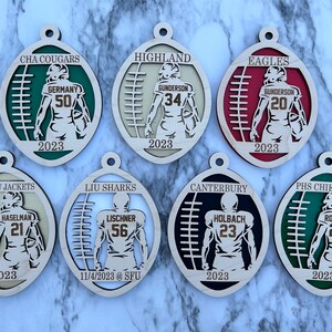 Op de afbeelding: Zes houten voetbalversieringen met teamnamen en spelersnamen. De versieringen zijn in verschillende kleuren geschilderd, waaronder groen, rood, zwart en wit. Elke versiering toont een voetballer in rugpositie met een voetbal in zijn handen. De versieringen zijn versierd met de teamnaam en de spelersnaam, evenals het jaar 2023.