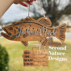 May include: Wooden fish-shaped sign with the name "Jackson Mark" in white script. The sign includes a smaller rectangular section with space to record date, time, weight, and length. The sign is held by a natural twine.