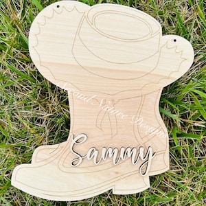 May include: A light-colored wooden cutout shaped like a cowboy boot with a hat resting on top. The piece features engraved outlines and the name Sammy in raised script lettering. Two small holes are visible at the top for hanging purposes.