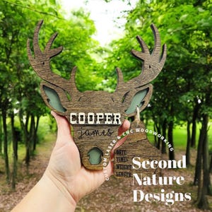 May include: A wooden deer head cutout with the name "COOPER james" engraved on it. The deer head is held in a hand and a wooden tag with the text "Second Nature Designs" is hanging from the bottom of the deer head.
