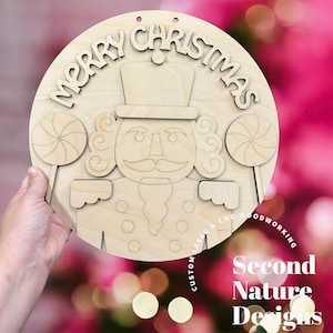 May include: A wooden Christmas decoration featuring a nutcracker design with the words "MERRY CHRISTMAS" at the top. The design includes two lollipops on either side of the nutcracker. The words "CUSTOM LASER & CNC WOODWORKING" and "Second Nature Designs" are also visible.