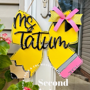 May include: A yellow wooden door hanger shaped like a pencil with the name "Ms. Tatum" in black script. The pencil design includes a pink eraser, gray metal, and a wooden tip. A decorative bow with pink and yellow accents is attached.