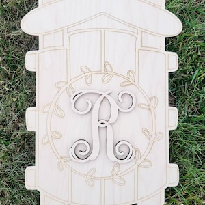 May include: A wooden sled-shaped sign with a large letter "R" in the center, surrounded by a wreath of leaves. The sign is unpainted, with etched details, and has a rustic, farmhouse aesthetic. Perfect for personalized home decor.