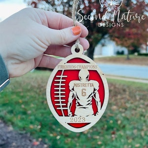 Op de afbeelding: Een houten voetbal ornament met een rood en wit ontwerp. De ornament heeft een voetballer in het midden met de tekst "Mistretta 6" op de achterkant van het shirt. De tekst "Firebirds Champions 2023" is ook op de ornament te vinden.