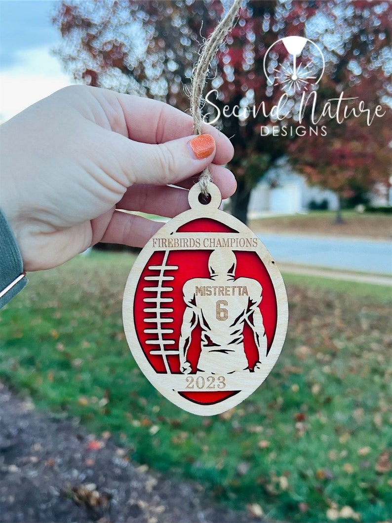 Op de afbeelding: Een houten voetbal ornament met een rood en wit ontwerp. De ornament heeft een voetballer in het midden met de tekst "Mistretta 6" op de achterkant van het shirt. De tekst "Firebirds Champions 2023" is ook op de ornament te vinden.