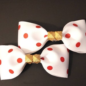 May include: Two white hair bows with red polka dots and gold accents. The bows are tied together with a gold connector.