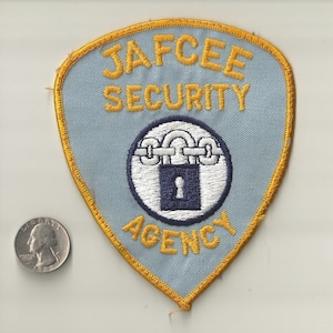 May include: A light blue JAFCEE Security Agency patch with gold trim. The patch features the text "JAFCEE SECURITY AGENCY" in gold lettering. A circular emblem in the center shows a lock and chain design. A U.S. quarter is next to the patch for size comparison.
