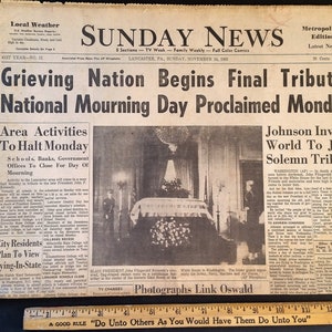 May include: A vintage newspaper, the Sunday News, from November 24, 1963, with the headline "Grieving Nation Begins Final Tribute." The front page features articles about the national mourning day and a photograph of a solemn event. A wooden ruler is at the bottom.