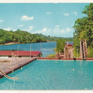 May include: A rectangular outdoor swimming pool with turquoise water, featuring a metal slide on the left and a tall diving structure on the right. A red-roofed building sits near a large blue lake surrounded by green trees under a blue sky.
