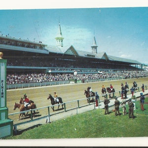 May include: A vintage postcard showing a horse racing track with a large grandstand filled with spectators. In the foreground, a green and white finish line post stands near the dirt track where jockeys on horseback are riding past.