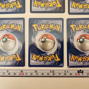 May include: Several Pok&eacute;mon trading cards are displayed, each featuring the Pok&eacute;mon logo and a Pok&eacute; Ball design on a blue background. The cards are arranged in rows, with a ruler placed below for measurement. The cards are in good condition.