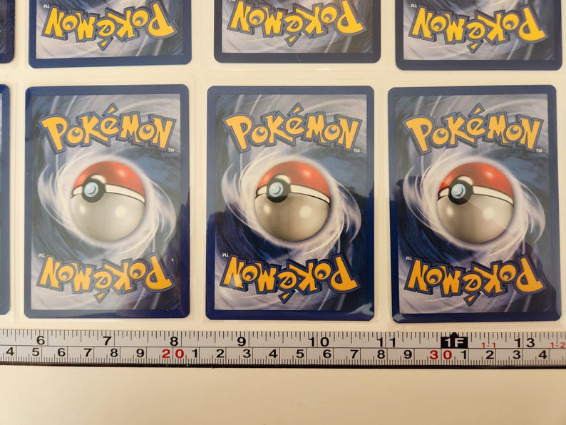 May include: Several Pok&eacute;mon trading cards are displayed, each featuring the Pok&eacute;mon logo and a Pok&eacute; Ball design on a blue background. The cards are arranged in rows, with a ruler placed below for measurement. The cards are in good condition.