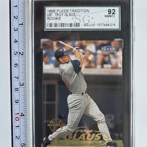 May include: A baseball card featuring Troy Glaus in action, from the 1998 Fleer Tradition series. The card is graded 92 NM/MT+ by SG, LLC. The card is in a black holder and includes the text "ROOKIE".