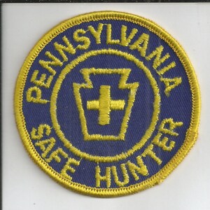 May include: A circular embroidered patch with a blue background and a yellow border. The text PENNSYLVANIA SAFE HUNTER is stitched in yellow around the perimeter. In the center, a yellow keystone shape contains a yellow cross symbol.